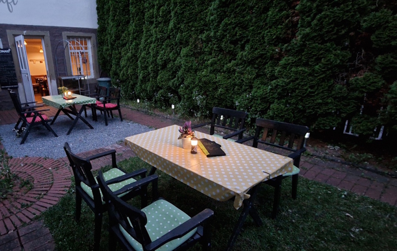 gartenwirtchaft/gartenoase bei dämmerung. 2 tische mit stühlen auf den tischen sind tischdecken, blumen, speisekarten und eine kleine lampe.
                im hintergrund sind hohe dichte bäume und der eingand in die gaststube. dort brennt das licht 