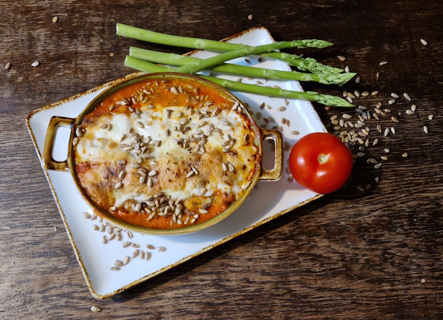 eine weiße quadratische auflaufform auf einem holzbrett. in der form sind gerollte kräuterpfannkuchen mit tomatensahnesoße und mit käse
           überbacken und gerösteten sonnenblumenkerne die auch als deko rechts neben der auflaufform liegne