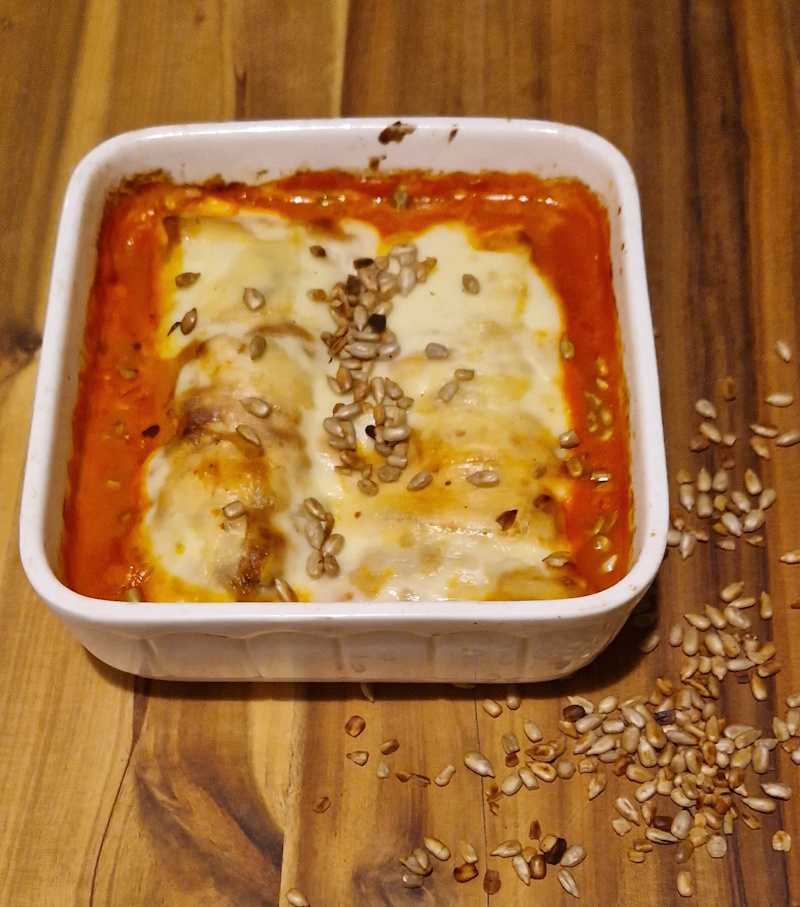 eine auflaufform mit 2 gerollten käuterpfannkuchen mit paprika-fetafüllung in einer tomatensahnesoße,
                mit käse überbacken. es liegen geröstete sonnenblumenkerne auf dem auflauf und als deko auf dem holzbrett