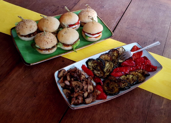 miniburger mit feta oder veggiepattie und pesto, mariniertes gemüße,eingelgten champignons,
                marinierte & gebackene spitzpaprika und zucchini auf 2 platten angerichtet auf einem alten holztisch
                mit gelbem tischläufer